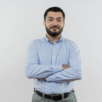 young bearded man in blue shirt looking at camera with crossed arms smiling confident standing over white background